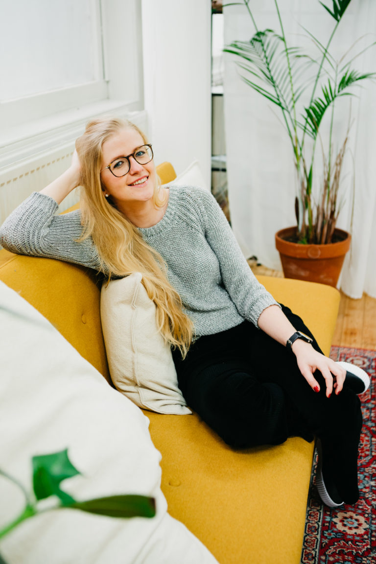 Lauren Van Noten zittend op een gele sofa in een huiselijke ruimte, met haar hand in haar haar en een glimlach.