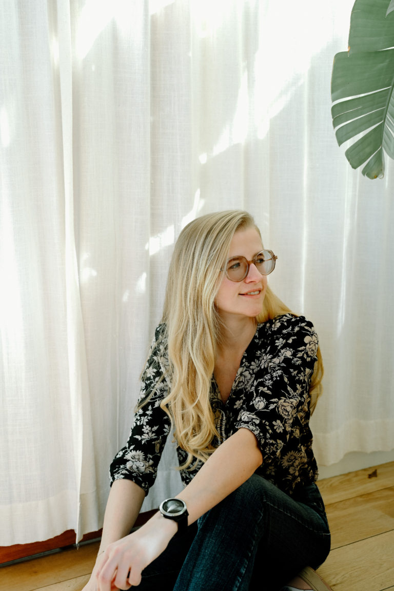 Lauren Van Noten zittend op de vloer voor lichte gordijnen, met een ontspannen blik en haar handen op haar knieën.