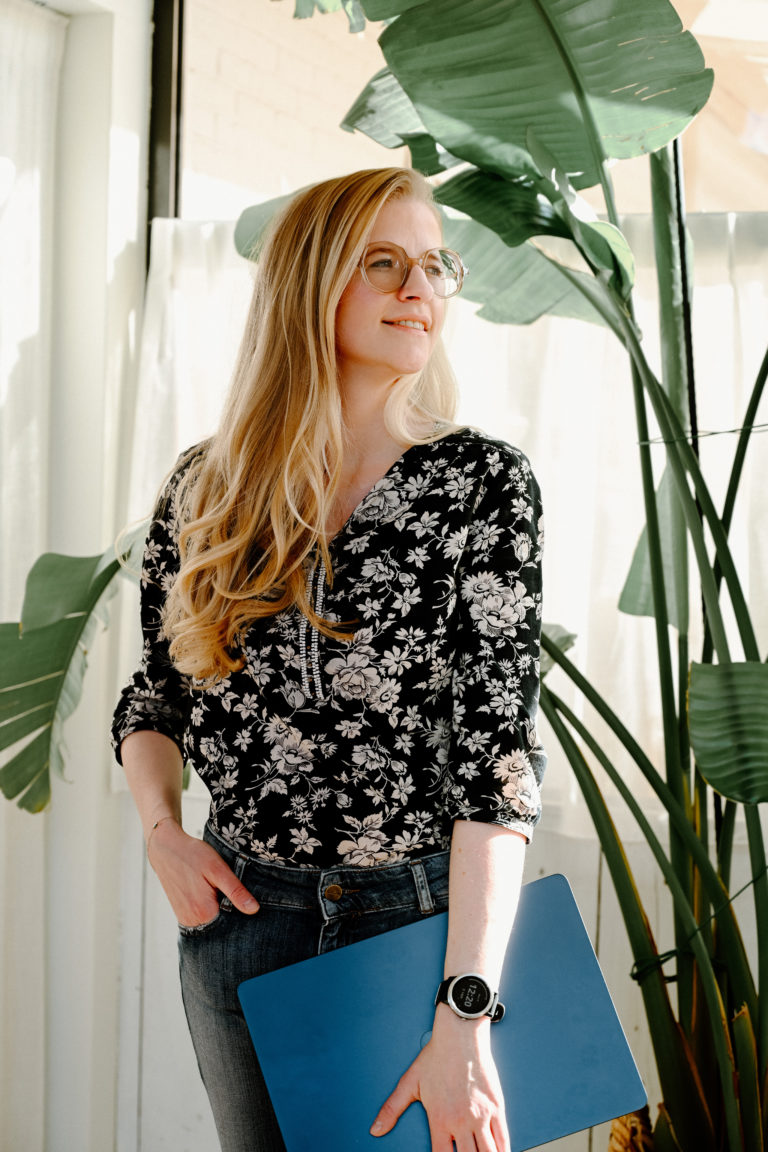 Lauren Van Noten staand in een lichte ruimte met grote planten, terwijl ze een blauwe laptop vasthoudt.