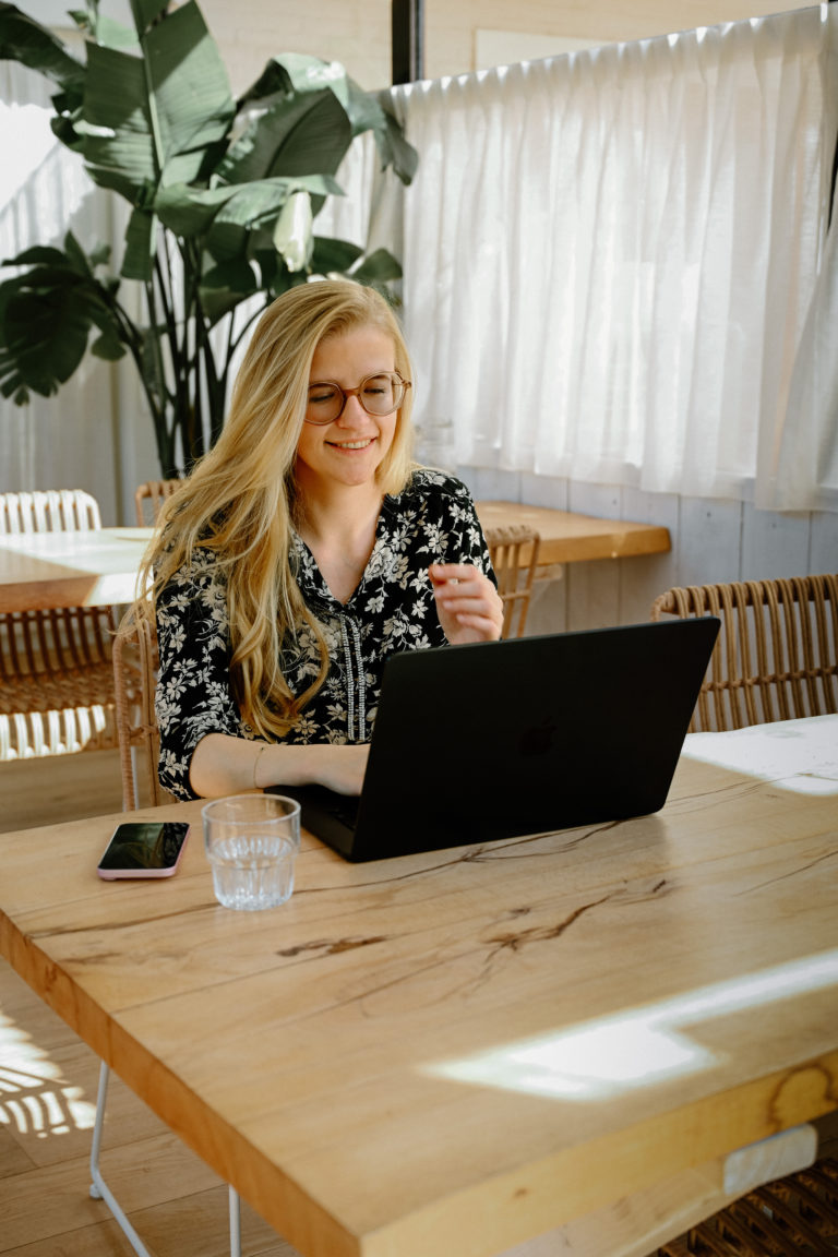 Lauren Van Noten, Notion expert, werkend op haar laptop aan een houten tafel in een lichte ruimte met grote planten op de achtergrond.