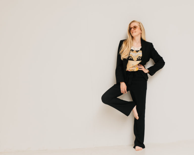 Lauren Van Noten staand tegen een lichte muur in een zwart pak, met één hand in haar zij en één voet omhoog tegen de muur.