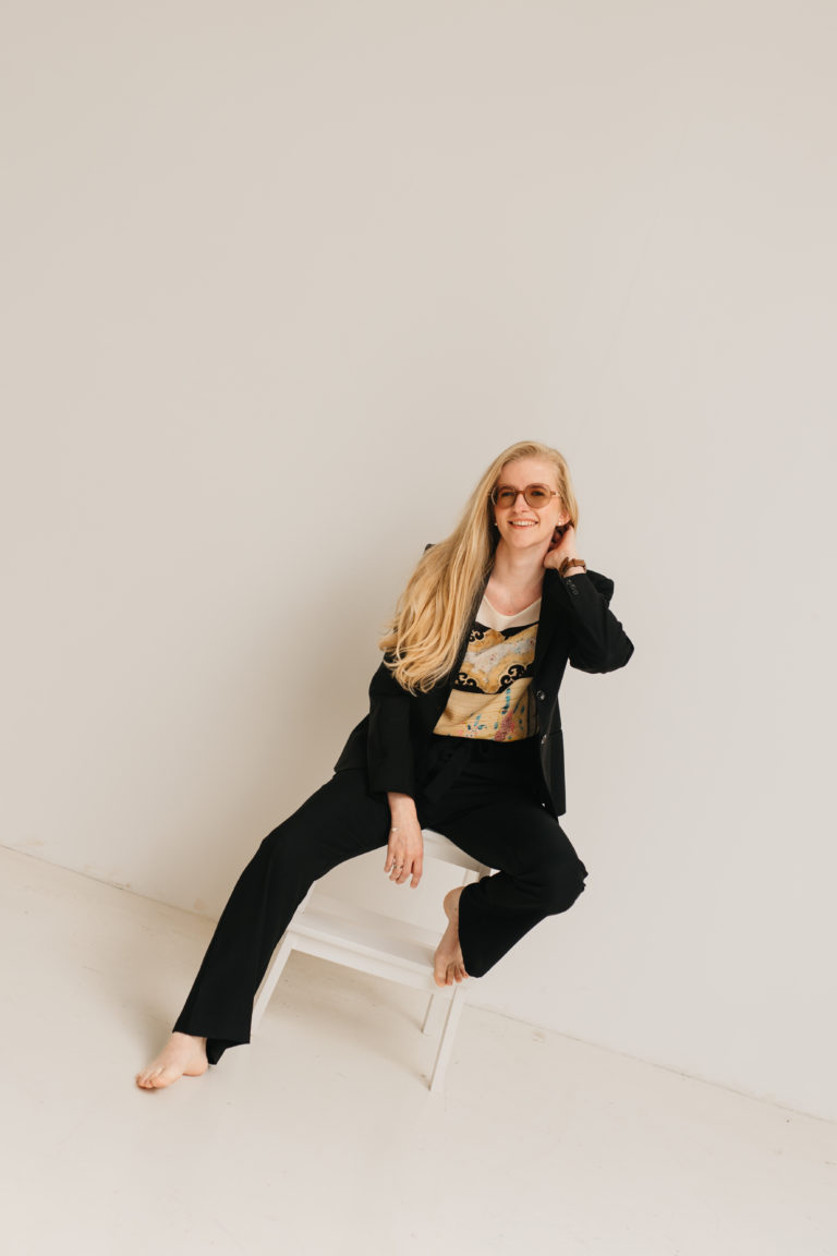 Lauren Van Noten zittend op een witte stoel in een minimalistische studio, gekleed in een zwart pak en met haar hand bij haar gezicht.