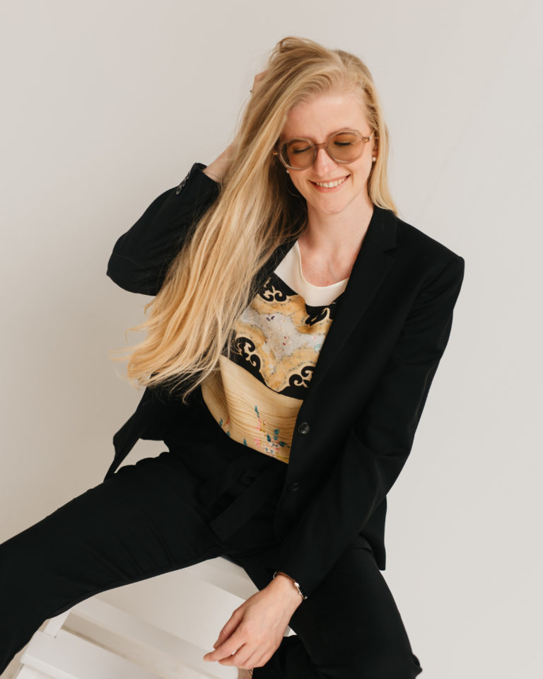 Lauren Van Noten zittend op een witte stoel in een lichte studio, gekleed in een zwart pak en met haar hand door haar haar.
