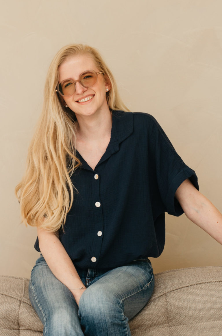 Lauren Van Noten die op de vloer zit tegen een lichte muur, met een donkerblauwe blouse en haar hand op haar knie.