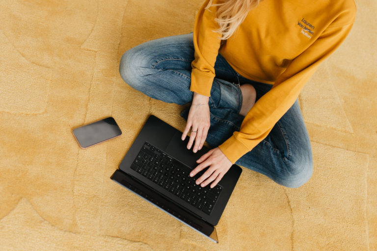Lauren Van Noten zittend op de vloer terwijl ze aan haar laptop werkt, met notitieboek en bril naast haar.
